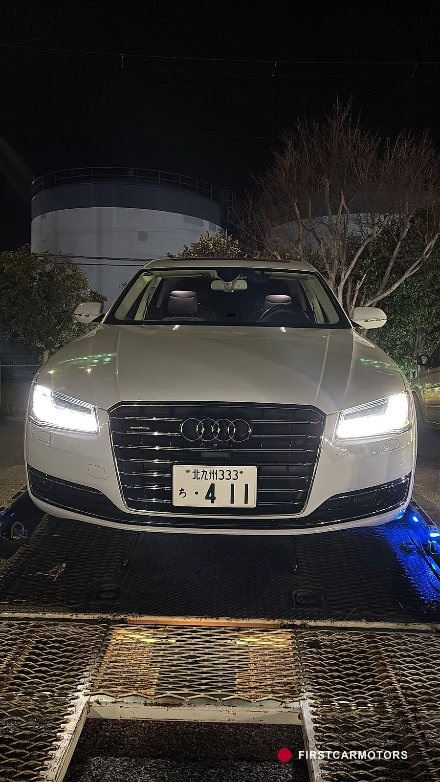 Ein weißer Audi A8 steht mit eingeschaltetem Licht auf der Ladefläche eines Flachbett-Transporters bei Nacht.